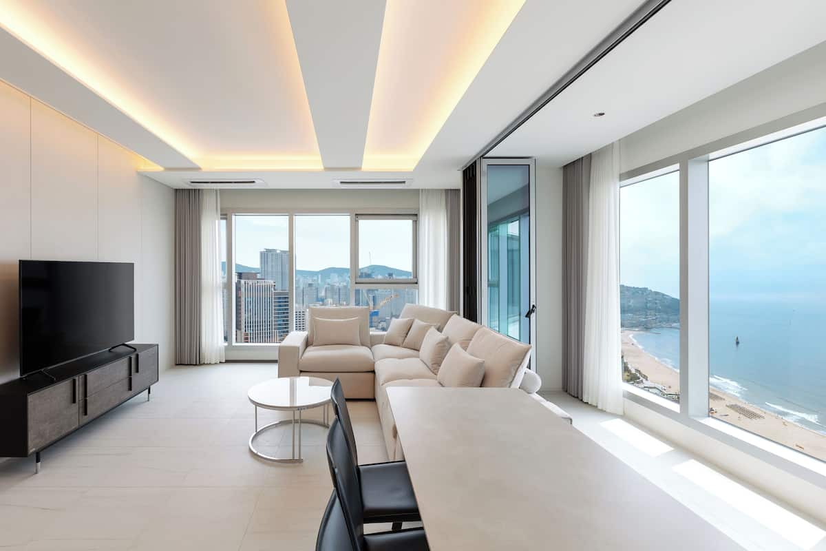 A modern hotel room with a large bed, a TV, a sofa, a dining table, and a view of the ocean.