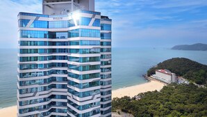 A high-rise hotel with a modern glass facade, located near a beach with a view of the sea and distant hills. - NewCZ Haeundae Residence (Busan)