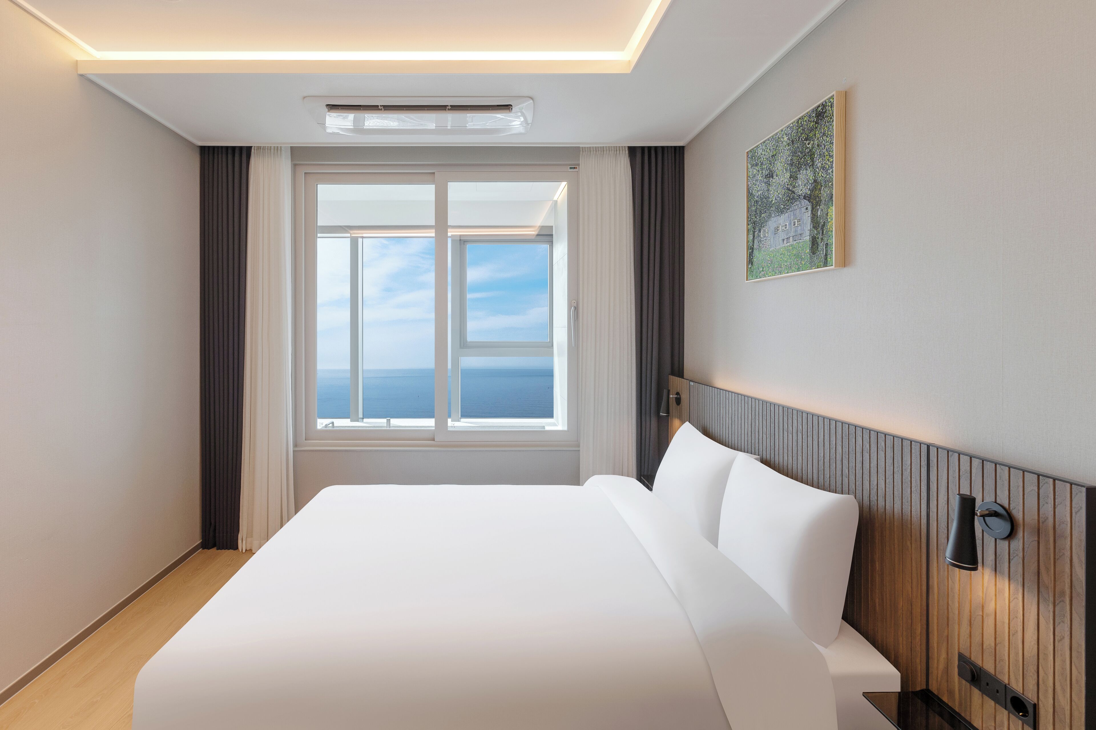 Une chambre d’hôtel moderne avec un grand lit, une vue sur la mer par la fenêtre et un tableau accroché au mur.