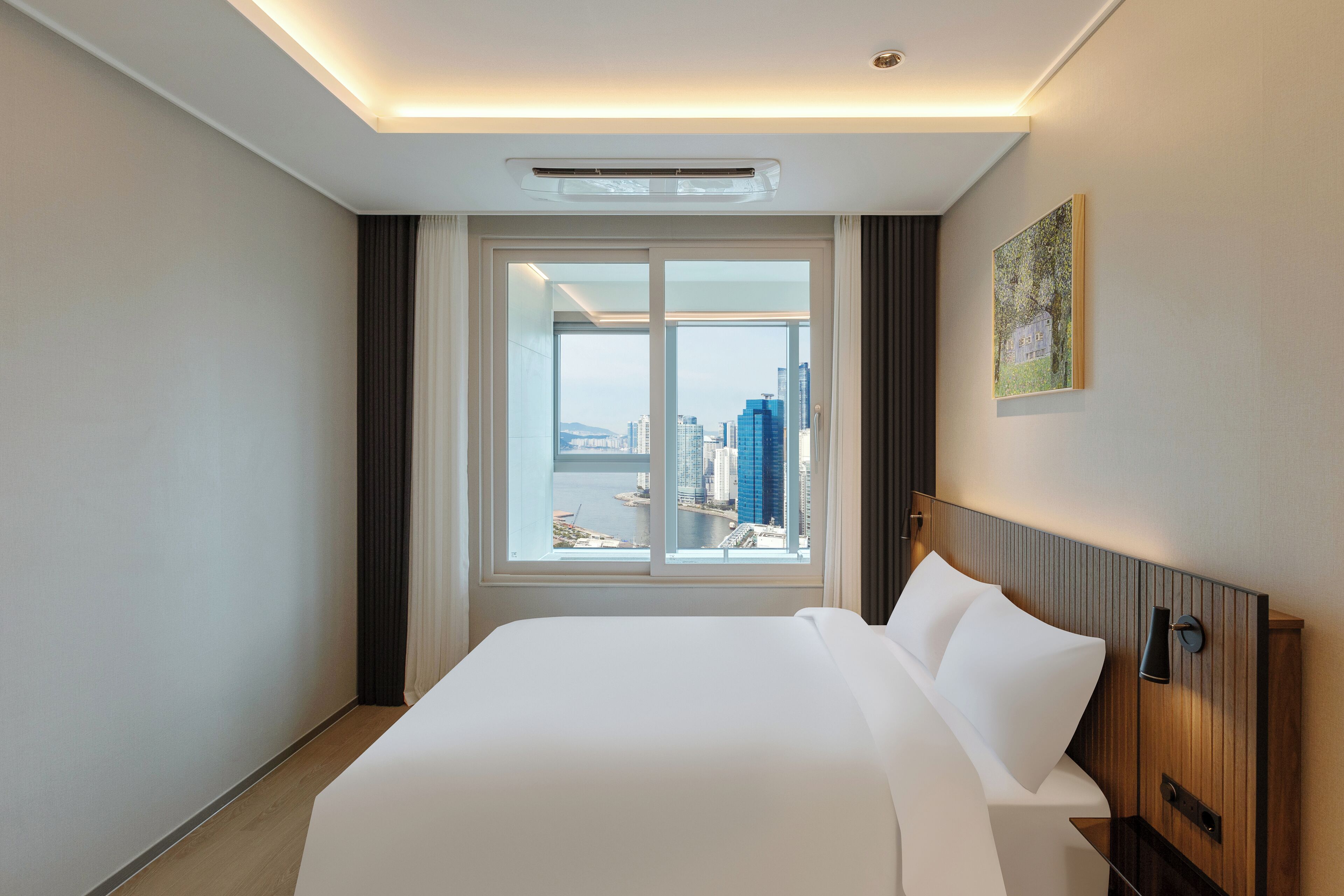 A hotel room with a large bed, a view of the city through a window, and a painting on the wall.