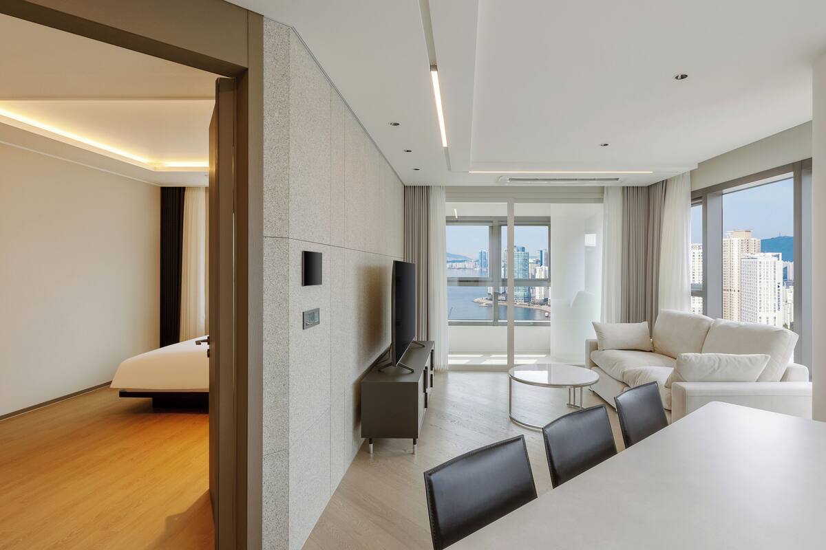 A modern hotel room with a large window, a sofa, a dining table, and a TV.