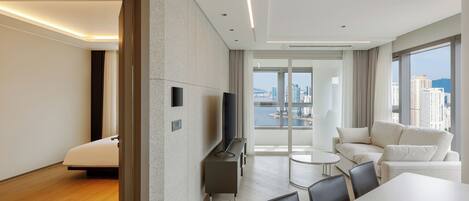 A modern hotel room with a large window, a sofa, a dining table, and a TV.