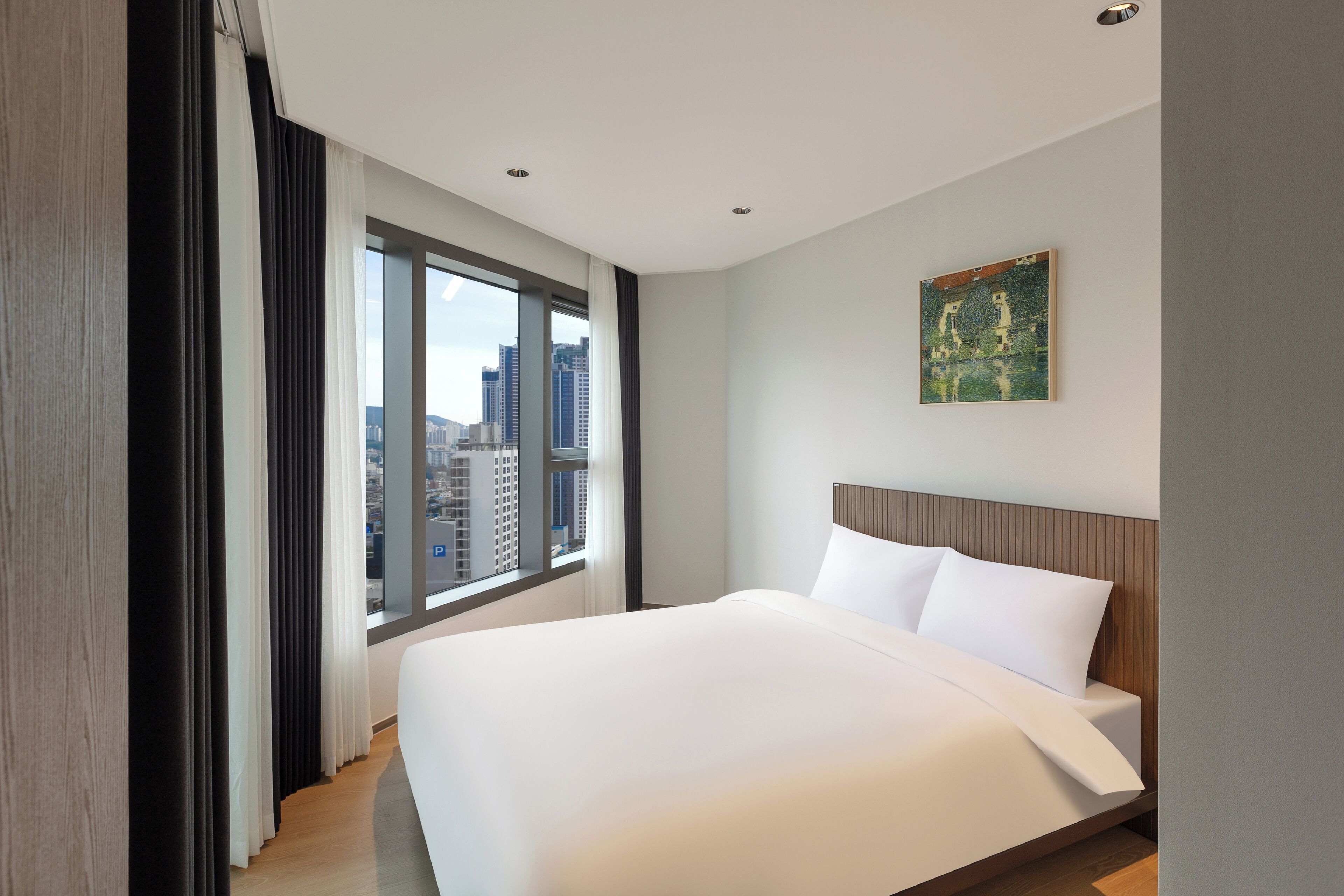 A modern hotel room with a large bed, a city view, and a painting on the wall.