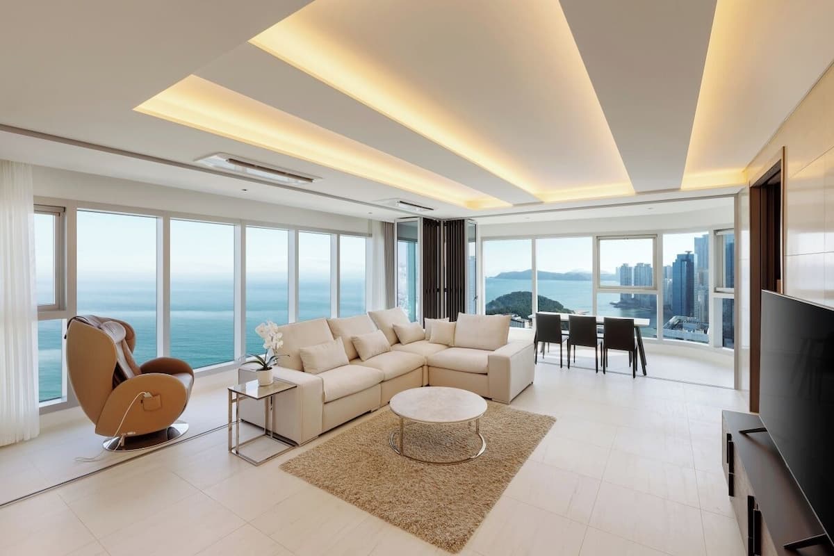 A modern living room with a large sofa, a coffee table, and a TV. There are large windows offering a view of the city and the sea.