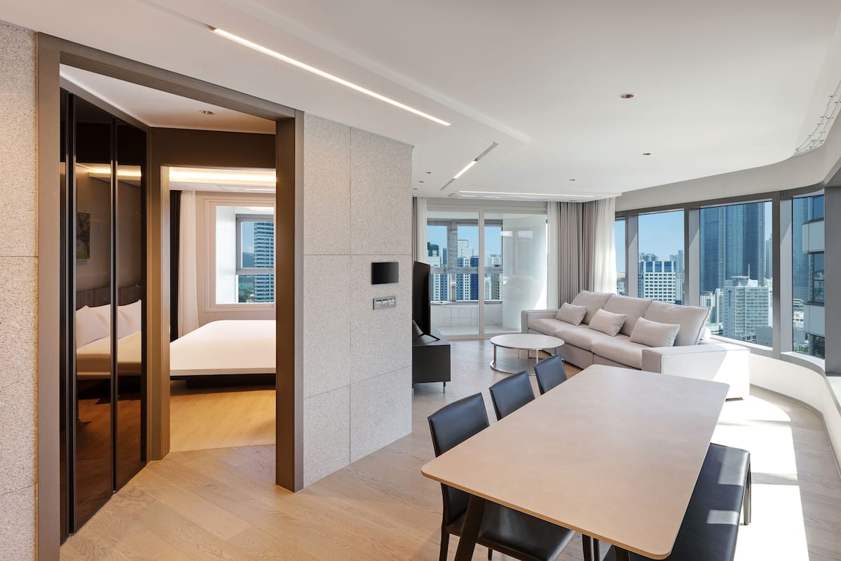 A modern hotel room with a bed, a dining area, a sofa, and a city view.