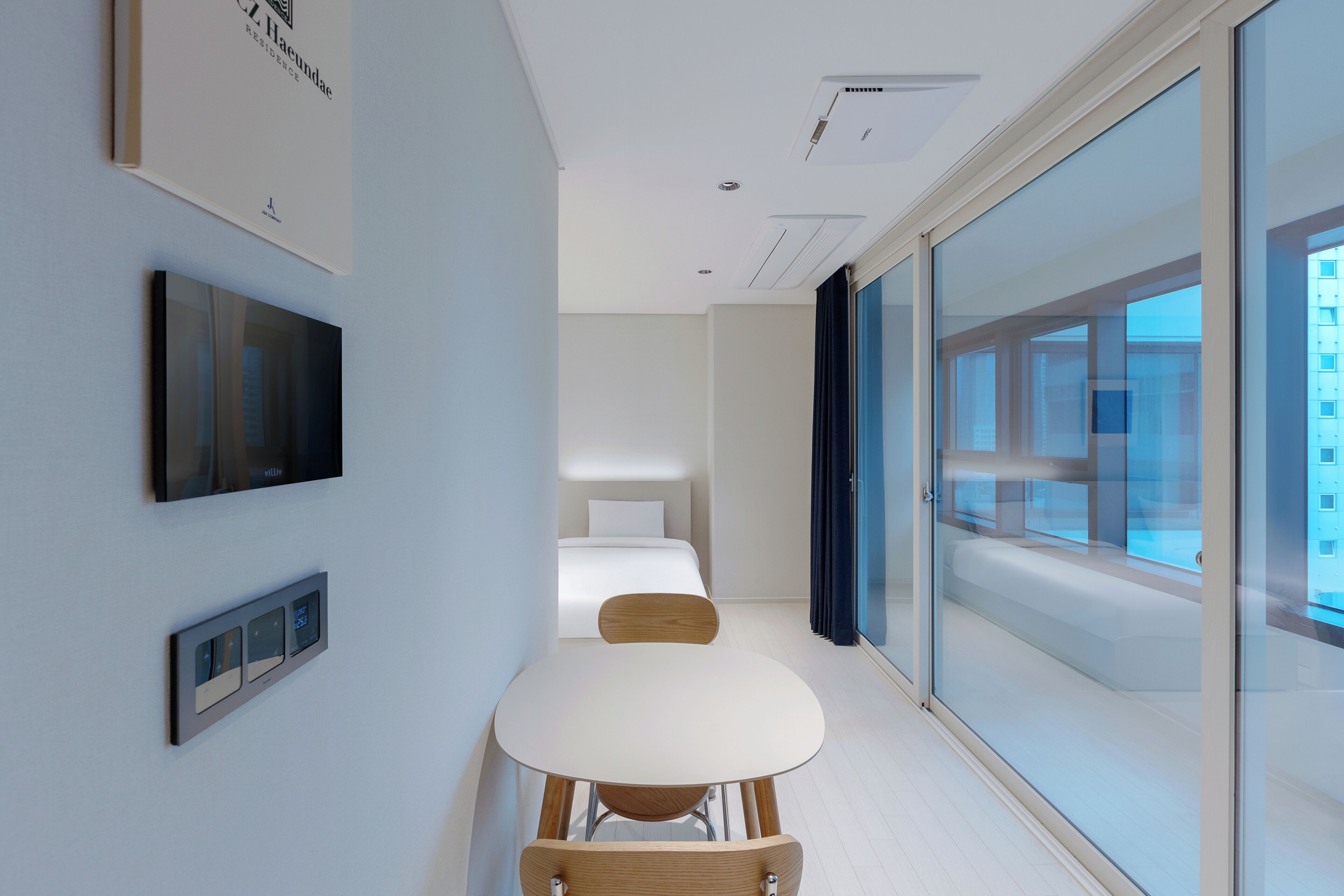 A modern hotel room with a bed, a TV, a digital thermostat, and a large window offering a city view.
