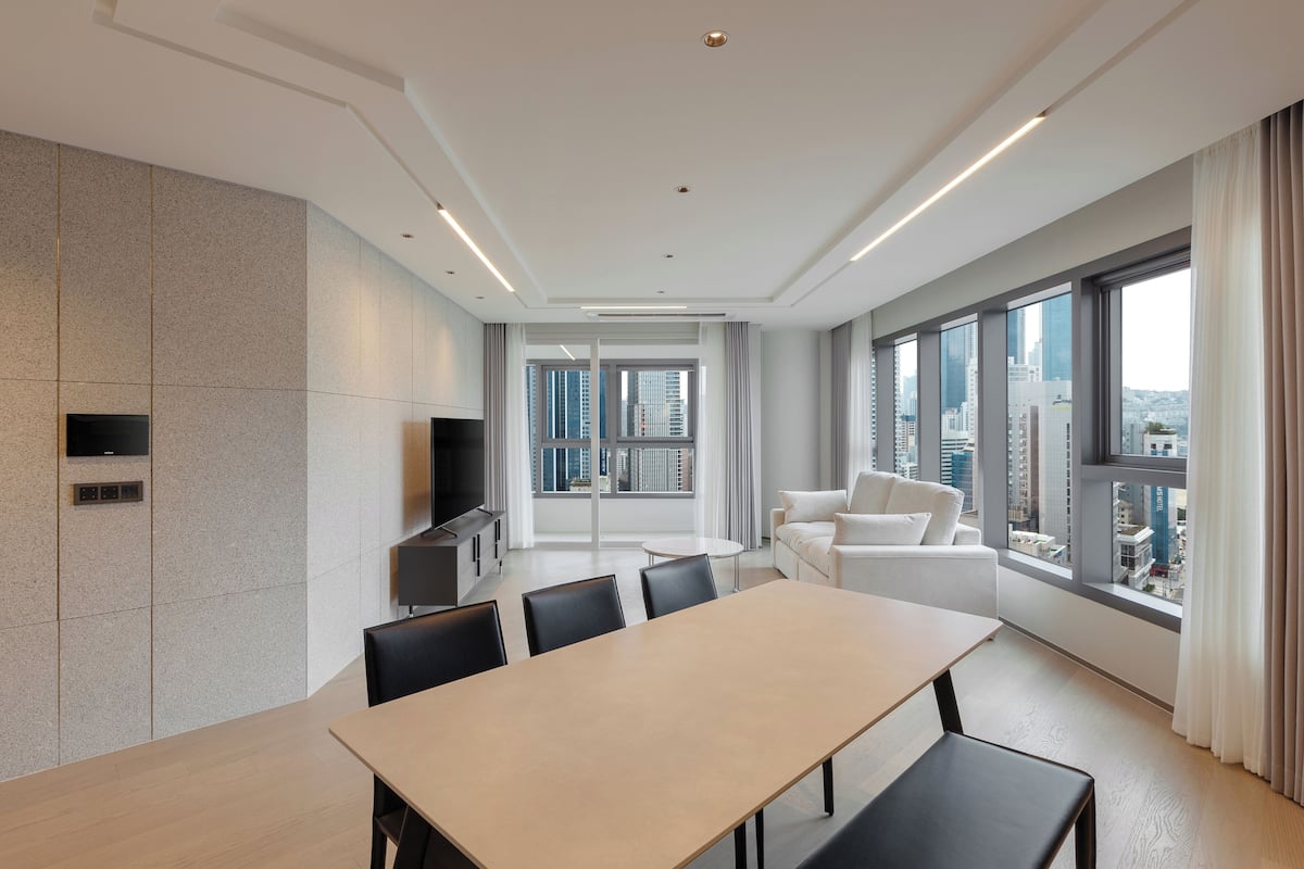 A modern hotel room with a large dining table, a sofa, and a TV.