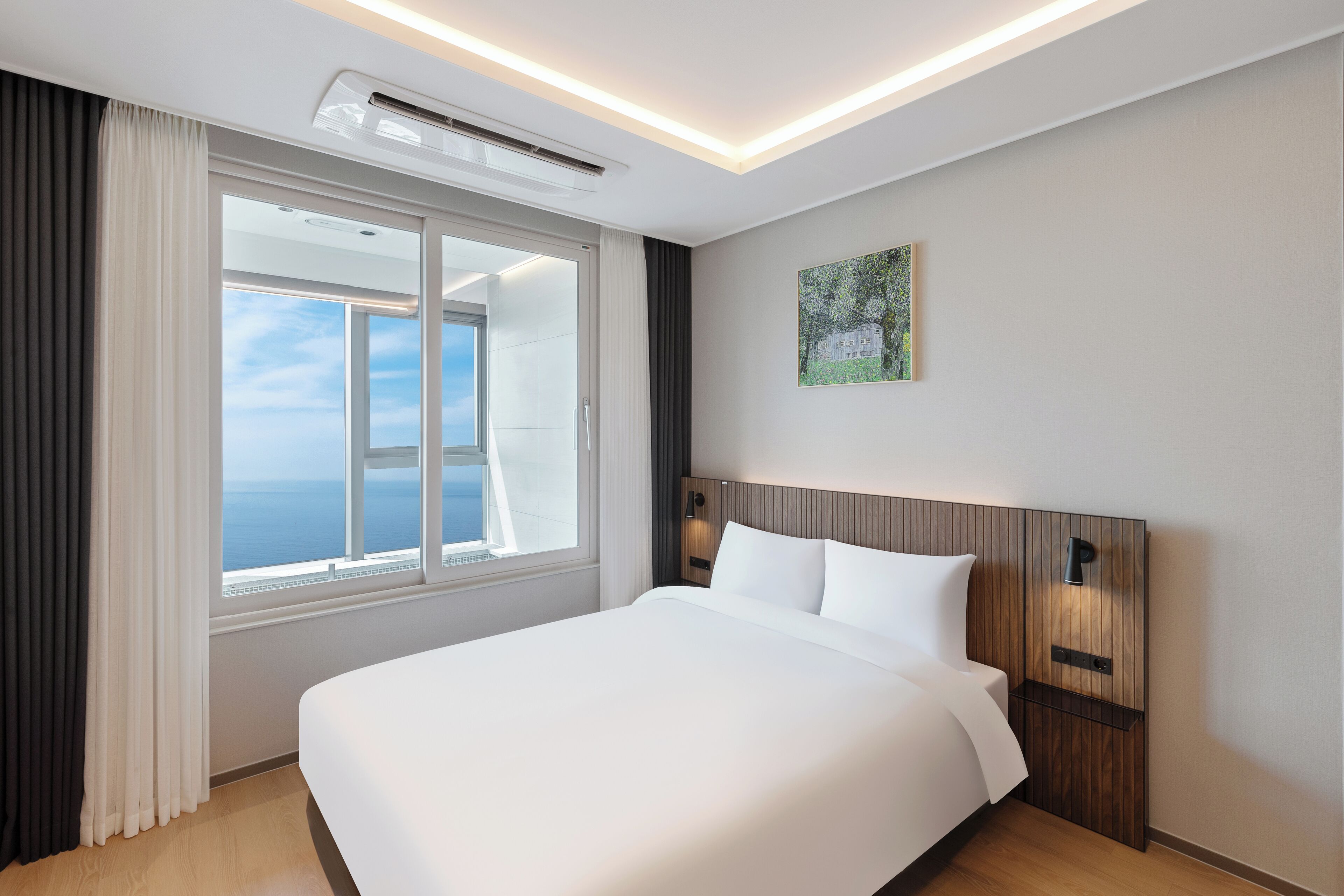 A hotel room with a large bed, a window with curtains, an air conditioning unit, a wall-mounted light, and a painting.