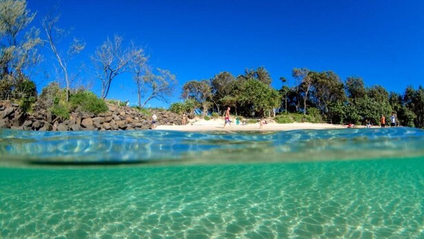 Een strand met helder turquoise water, een rotsachtige kustlijn en bomen op de achtergrond.