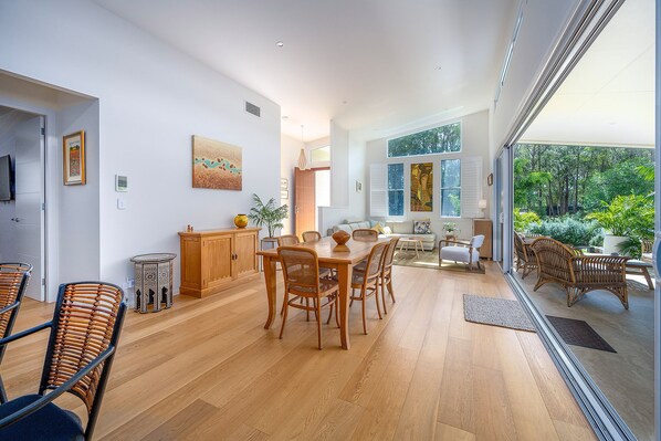 A spacious dining area with a wooden table and chairs, a small cabinet, and a view of greenery through large windows. - Brunswick Lux - Close to Creek & Beach (Brunswick Heads)