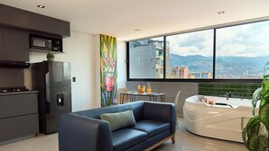 A modern living space with a sofa, a kitchen area, a dining table with chairs, a large window with a city view, and a bathtub. - Jacuzzi Retreat | Cozy Aparta-Suite | Rosa Negra (Medellín)