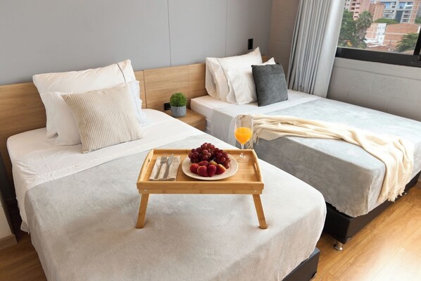 Habitación de hotel con dos camas, una bandeja de madera con fruta y jugo, y vista a edificios a través de la ventana.