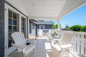Een overdekte veranda met witte Adirondack-stoelen en een tafel, een klein blauw huis op de achtergrond, en een strakblauwe lucht.