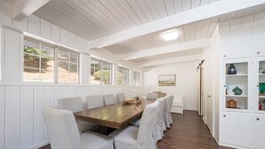 A dining area with a long wooden table and white upholstered chairs, white paneled walls, and a view through large windows. - Bohemian Oasis on Vine (Paso Robles)