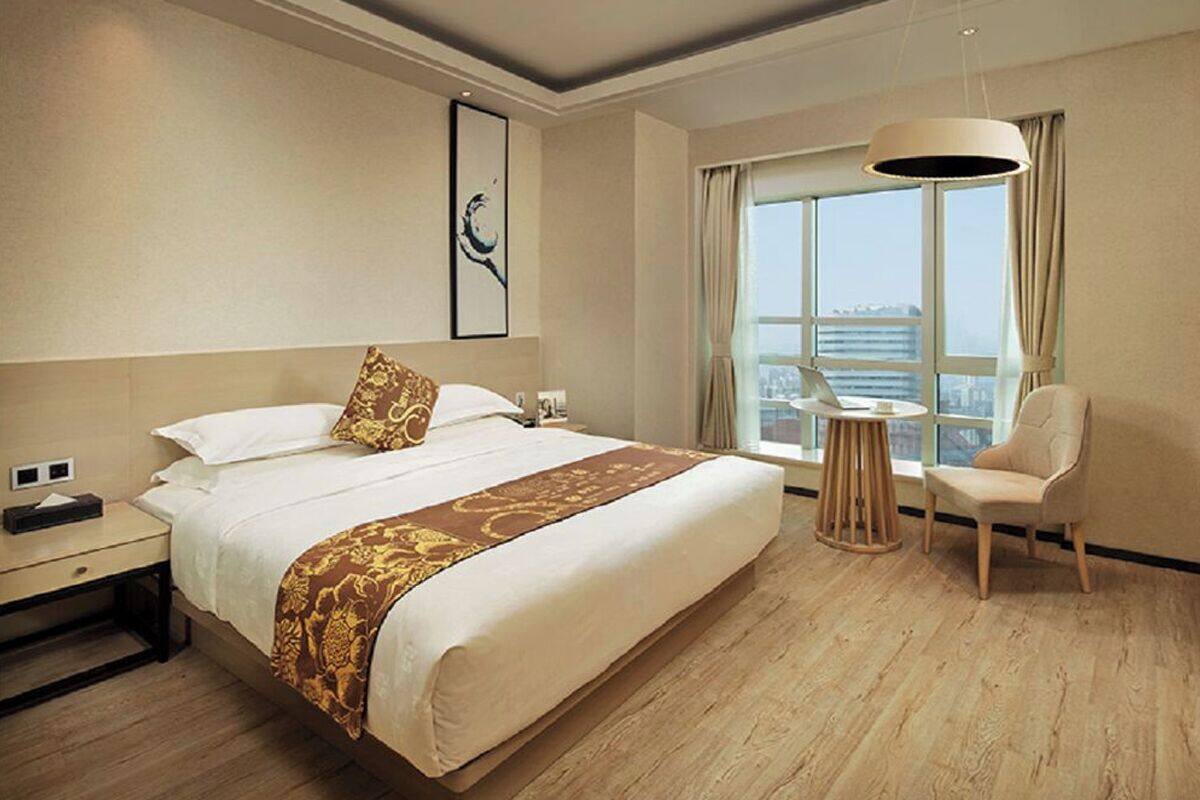 A hotel room with a large bed, a desk, a chair, a TV, and a large window with a view of the city.
