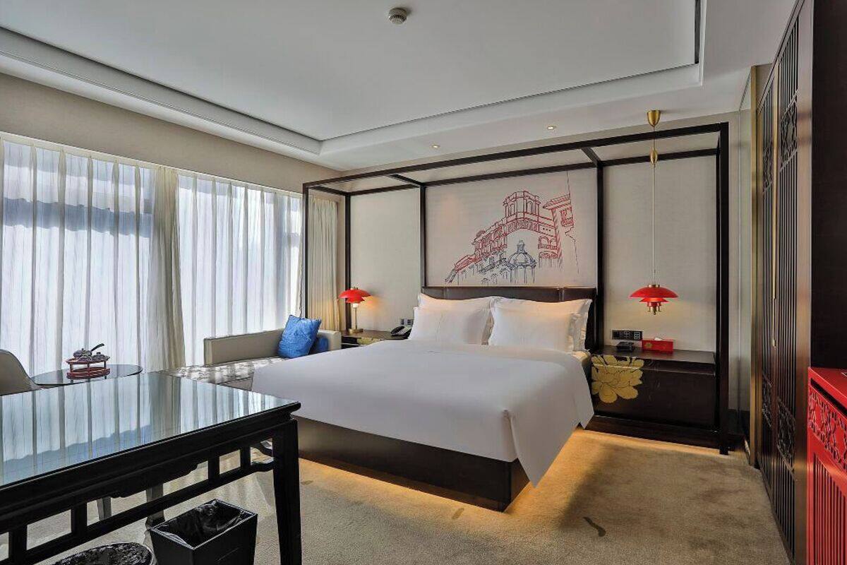 A hotel room with a large bed, bedside tables, a desk, and a view of the cityscape through the window.
