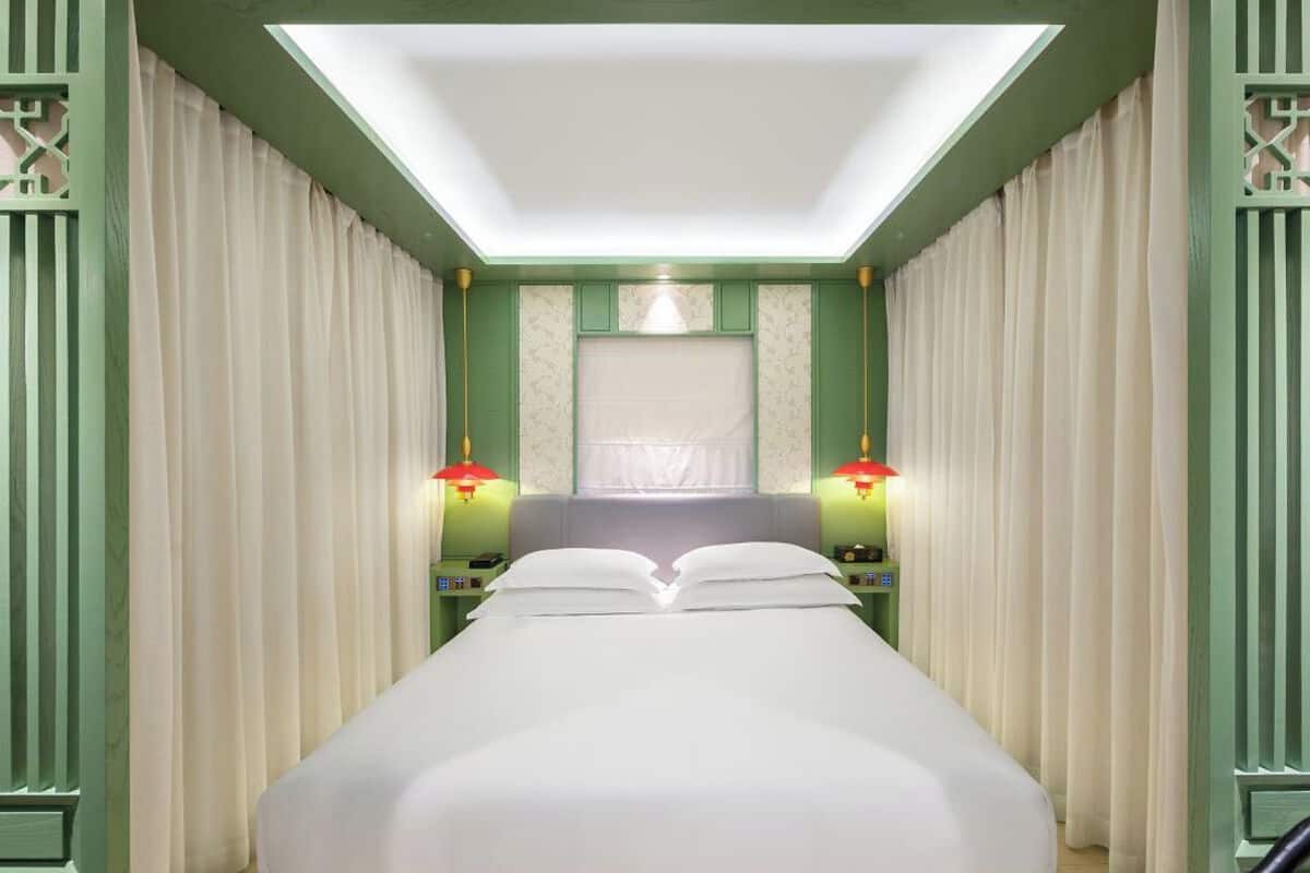 A hotel room with a bed centered under a skylight, flanked by green walls and white curtains.