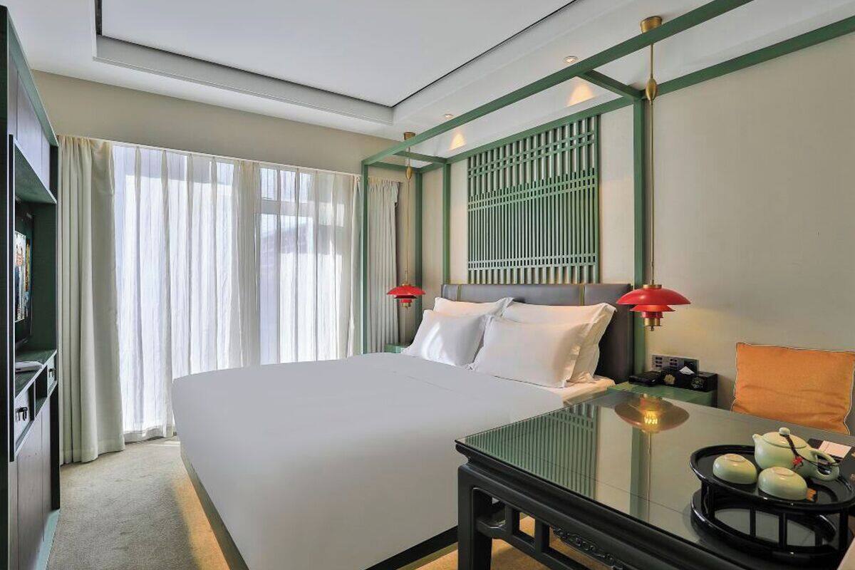 A hotel room with a large bed, a glass coffee table, a red lamp, and a view of the outdoors.