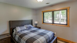 A bedroom with a bed, nightstand, lamp, and a window with a view of trees. - Cabin w/Bunk on Whitefish Chain! (Crosslake)