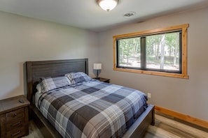 A bedroom with a bed, nightstand, lamp, and a window with a view of trees. - Cabin w/Bunk on Whitefish Chain! (Crosslake)