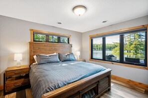 Ein Schlafzimmer mit einem hölzernen Kopfteil, einem großen Fenster mit Blick auf Bäume, einem Bett mit blauer Bettwäsche und zwei Nachttischen mit Lampen.