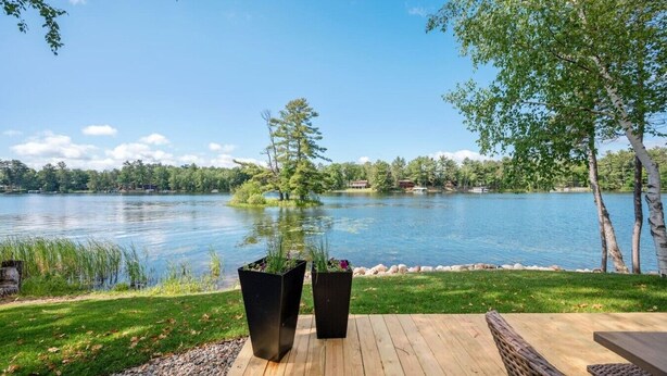 A lakeside deck with two planters, a wooden deck, and a view of a lake and distant trees. - Cabin w/Bunk on Whitefish Chain! (Crosslake)