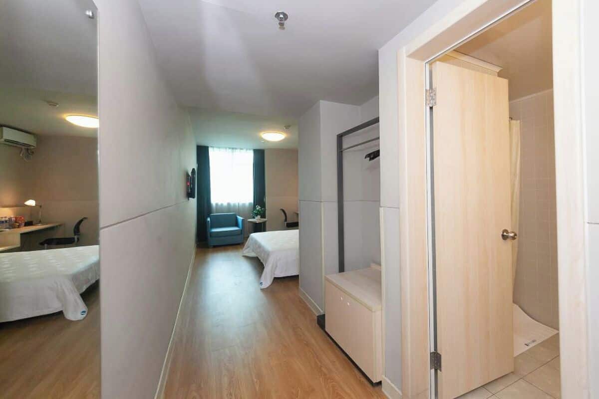 A hotel room with a wooden floor, a bed, a desk, a chair, a small table, a mirror, and a door leading to a bathroom.