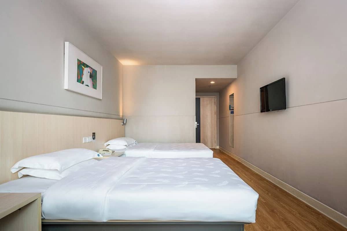 A hotel room with two beds, a bedside table, a TV mounted on the wall, and a framed picture on the wall.