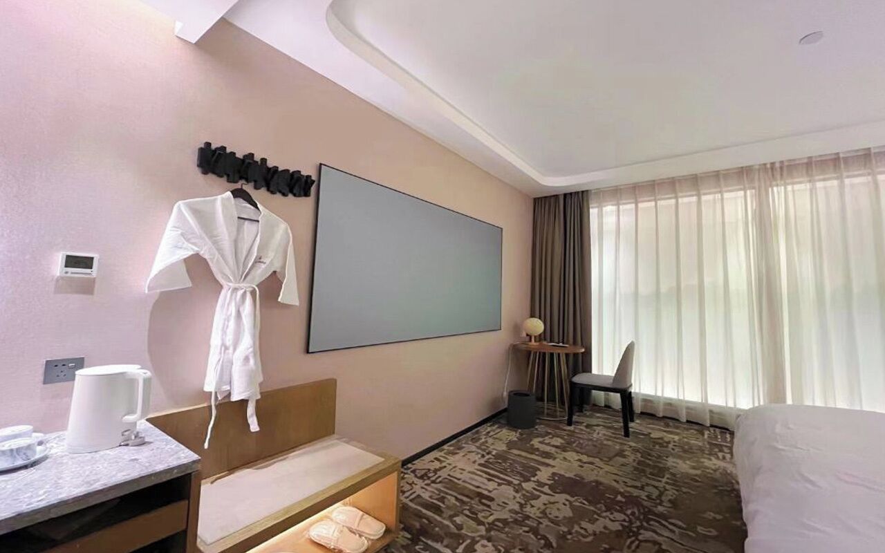 A hotel room with a bed, a desk with a chair, a robe hanging on the wall, a large window with curtains, and a flat-screen TV.