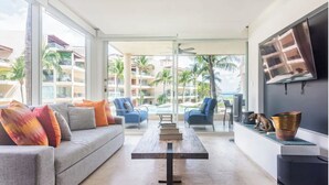 A modern living room with a grey sofa, blue armchairs, and a wooden coffee table. A TV is mounted on the wall, and a large window offers a view of a pool and palm trees.