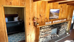 Un interior de madera con chimenea de piedra que dice "BLUE MOUNTAIN LAKE", una cama y vistas de montañas a través de una ventana.