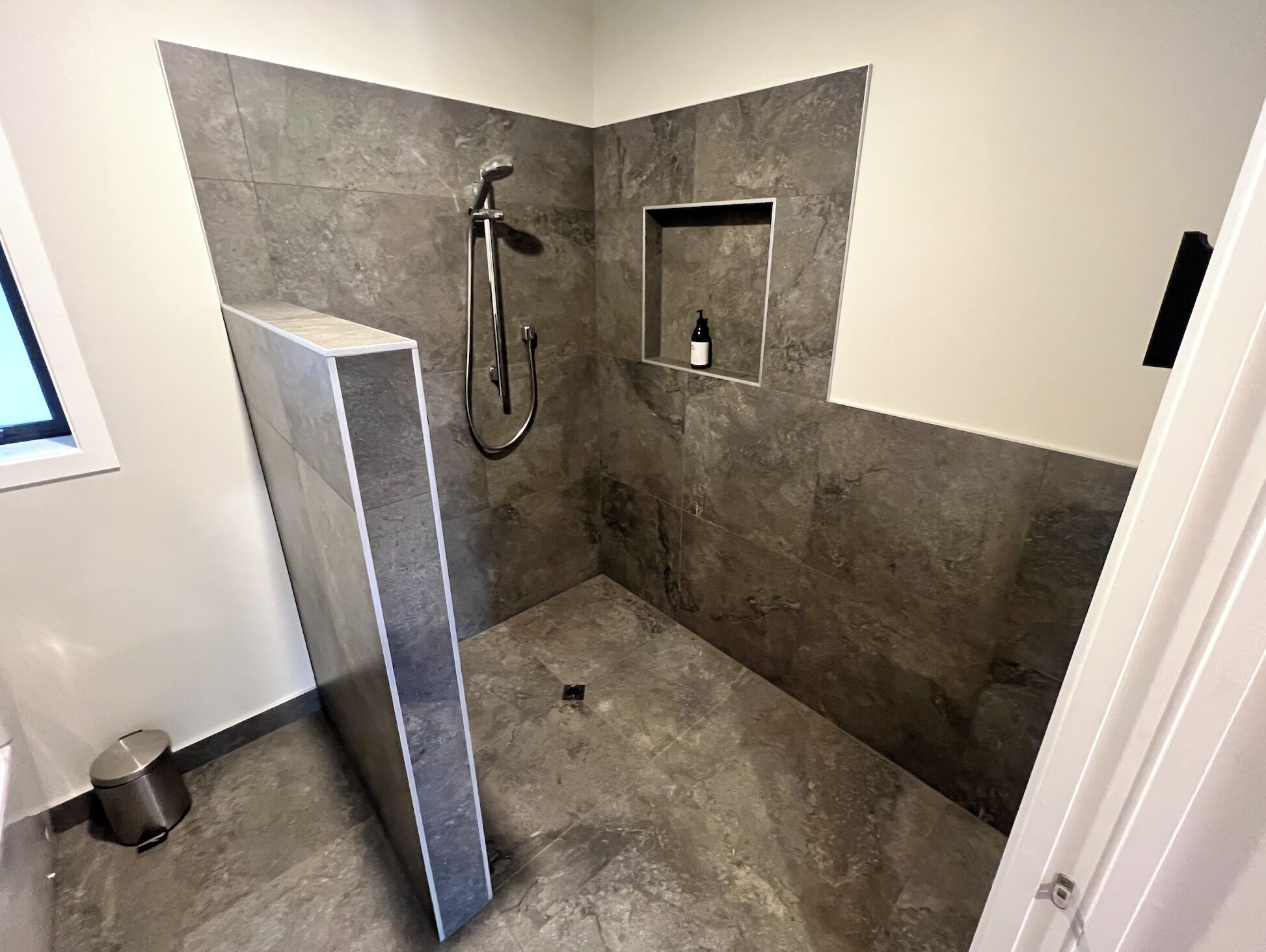 A modern bathroom with a shower, a built-in shelf, and a trash can.