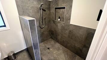 A modern bathroom with a shower, a built-in shelf, and a trash can.