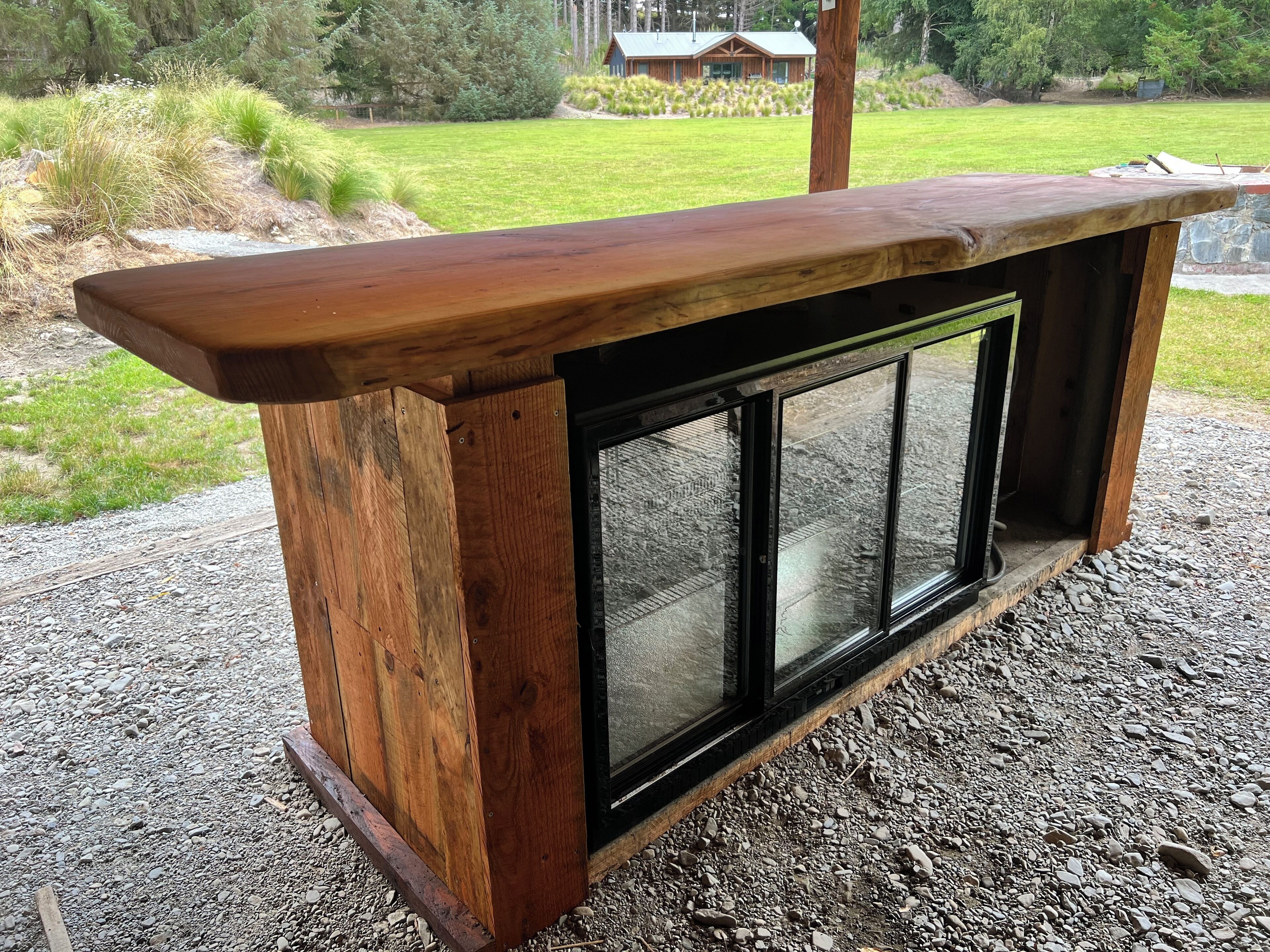 A wooden bench with a glass enclosure, set on a gravel surface with a grassy area and trees in the background.