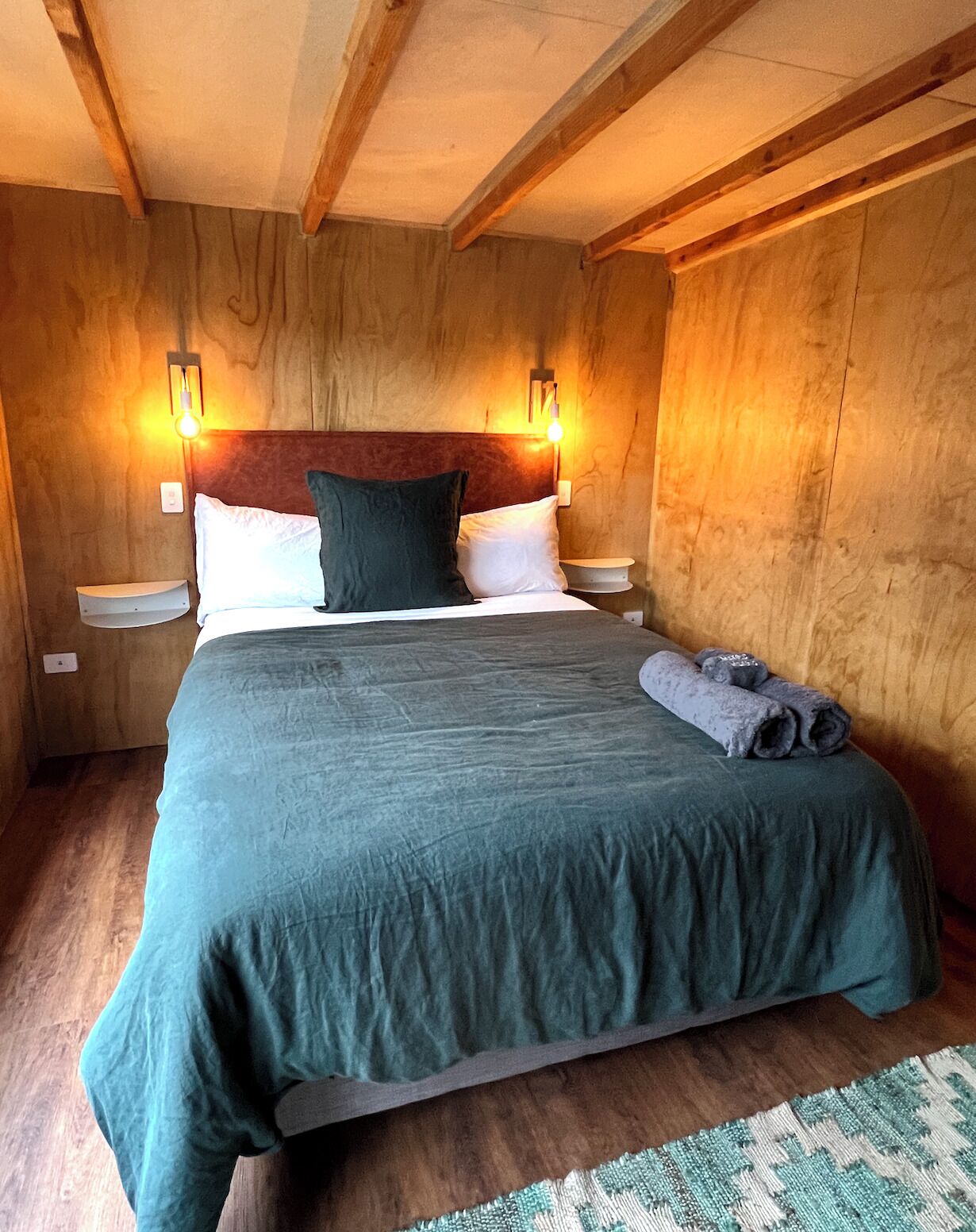 A wooden-paneled room with a bed, two bedside tables, and wall-mounted lights.