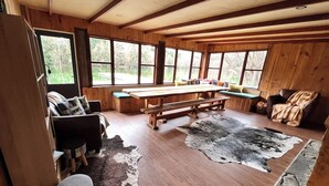 Interior rústico de una cabaña con paredes de madera, ventanas grandes, una mesa de comedor de madera, bancos y una alfombra de piel.