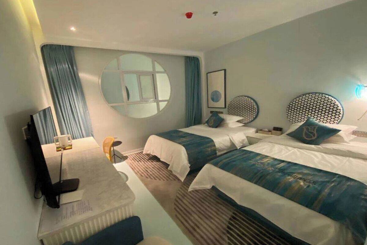 A hotel room with two beds, a TV, a desk, and a large window with curtains.