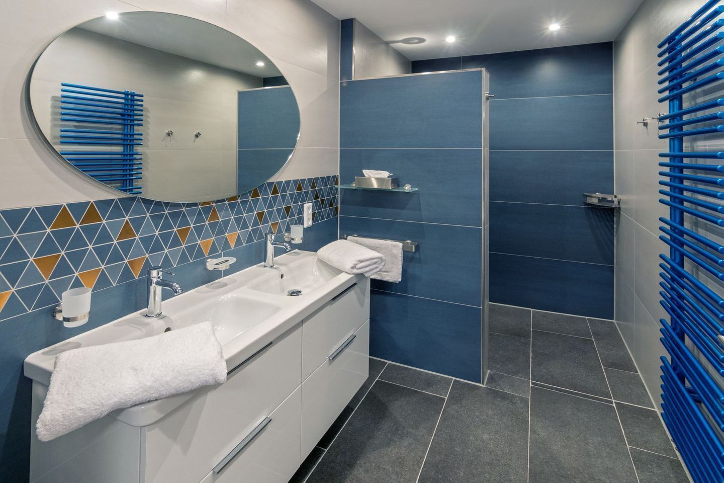 Een moderne badkamer met een witte wastafel, een ronde spiegel en een douchegedeelte met blauwe tegels.