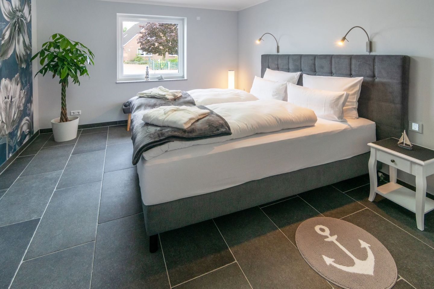 Un dormitorio moderno con una cama grande, una mesita de noche con lámpara, una planta en maceta y vistas al exterior a través de una ventana.