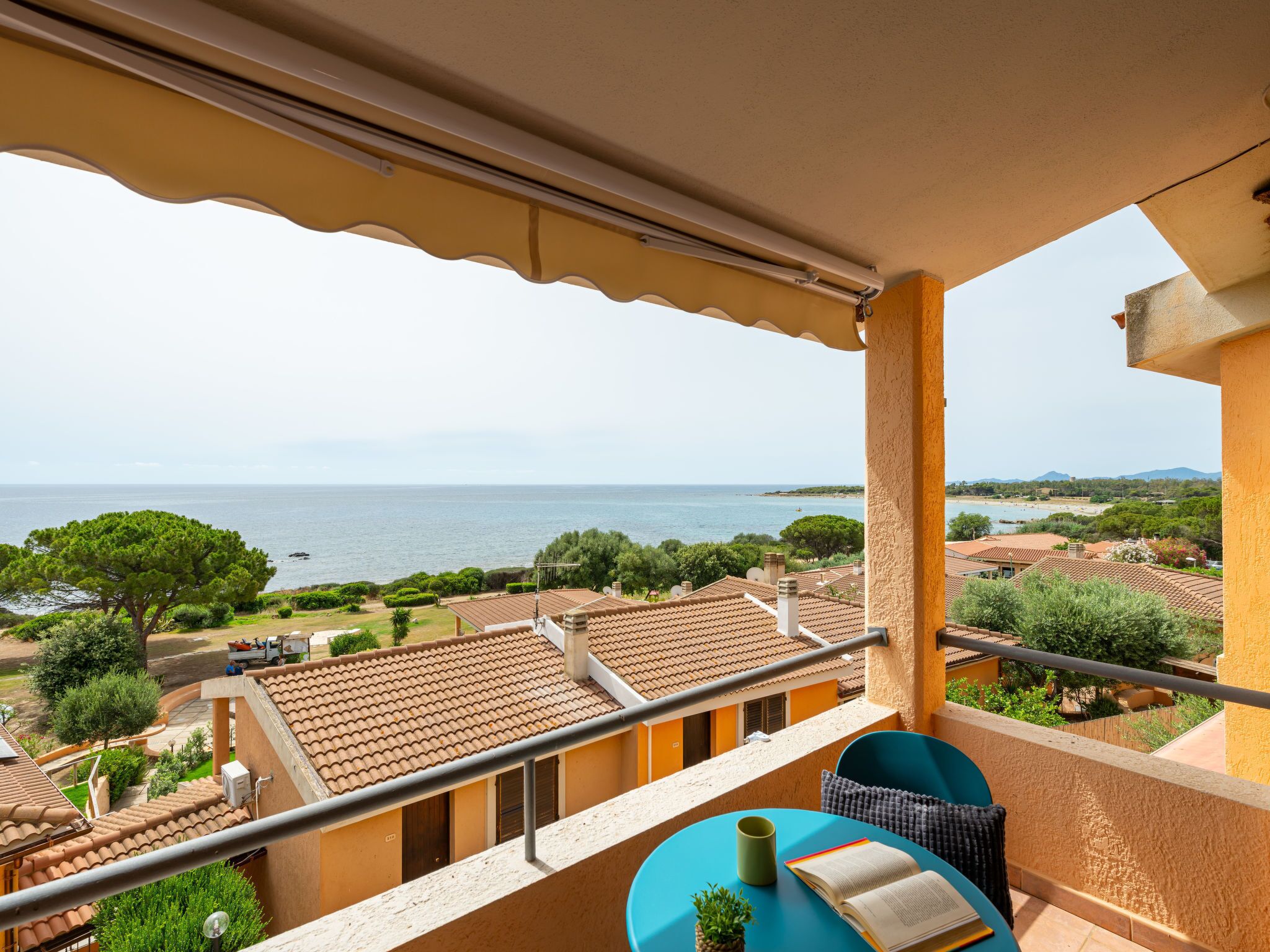 A balcony with a view of the sea, a table with a book and a plant, and a chair.