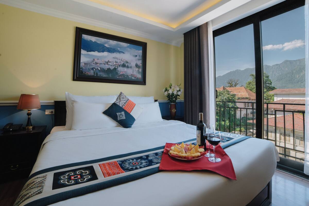Ein Hotelzimmer mit einem großen Bett, einem Gemälde einer Berglandschaft, einem Nachttisch mit Lampe und Blick auf Gebäude vom Balkon aus.