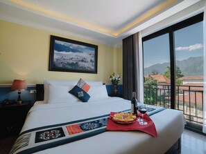 A hotel room with a large bed, a painting of a mountain landscape, a nightstand with a lamp, and a view of buildings through a balcony. - Ladybird Sapa Hotel by Minova (Sa Pa)