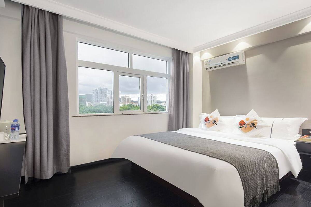 Habitación de hotel con una cama grande, una ventana con vistas a la ciudad, una mesita de noche con una botella de agua y un equipo de aire acondicionado.