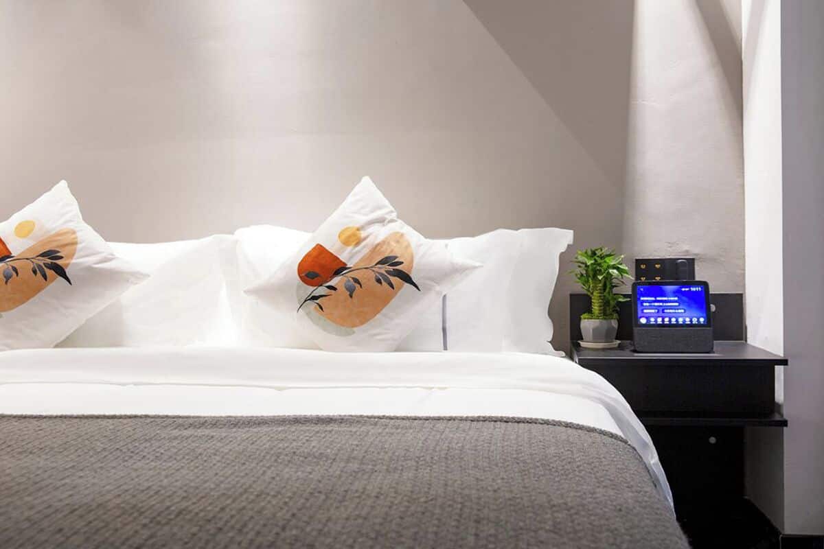 Una cama bien hecha con ropa blanca y almohadillas decorativas, una mesita de noche con un reloj digital y una planta en maceta.