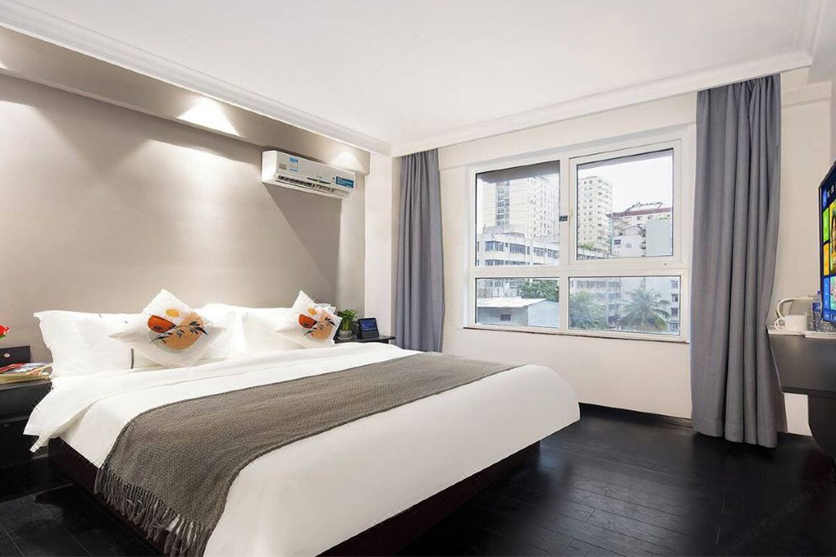 Una habitación de hotel con una cama grande, una mesita de noche con una lámpara, un equipo de aire acondicionado, una ventana con vistas a edificios y una cafetera sobre una mesa.