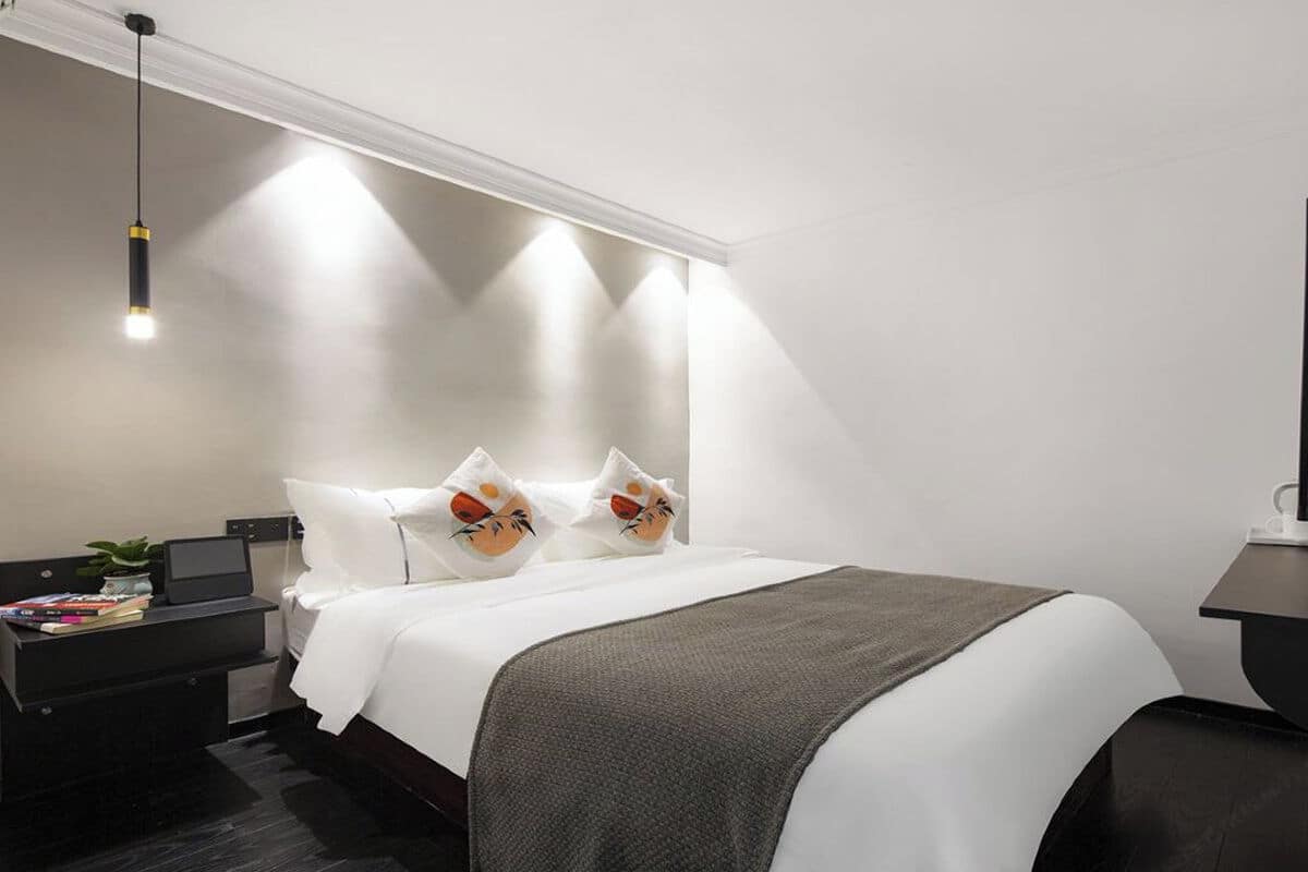Una habitación de hotel moderna con una cama bien hecha, una mesita de noche con una computadora portátil, una planta pequeña y un televisor de pantalla plana.