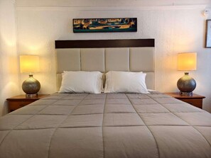 Ein ordentlich bezogenes Bett mit einer gesteppten Decke, zwei weißen Kissen und einem Kopfteil mit dunkler, horizontaler Zierleiste. Auf den Nachttischen aus Holz stehen je eine Nachttischlampe mit gemusterten Fußleisten. Ein Gemälde hängt über dem Bett.