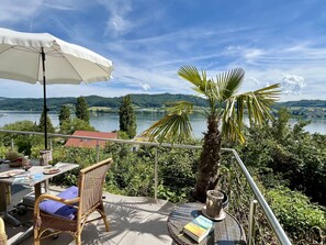 Un balcone con mobili in vimini, un ombrellone bianco e vista su un lago con alberi e colline sullo sfondo.