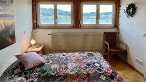 A bedroom with a large window, a radiator, a bed with a colorful quilt, a chair, and a wall-mounted wreath. - Seepanorama by Interhome (Öhningen)
