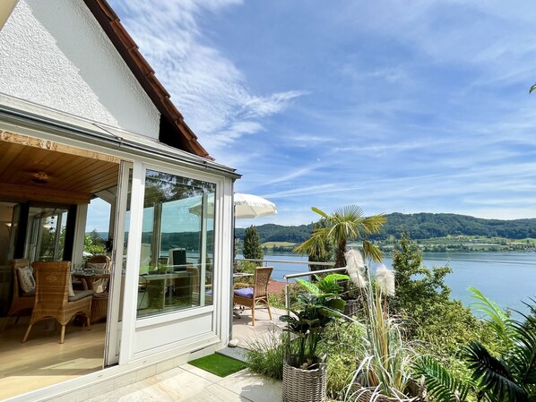 Una casa moderna con una porta in vetro che dà su un'area pranzo con mobili in vimini e vista su un lago.
